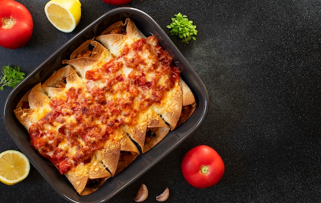 Mexikanische Enchiladas mit Huhn, Gemüse, Mais, Bohnen, Tomatensauce und Käse. Im Backblech serviert. Mexikanische Nahrung. Lateinamerikanische Küche. Schwarzer Hintergrund, Draufsicht, Kopierraum