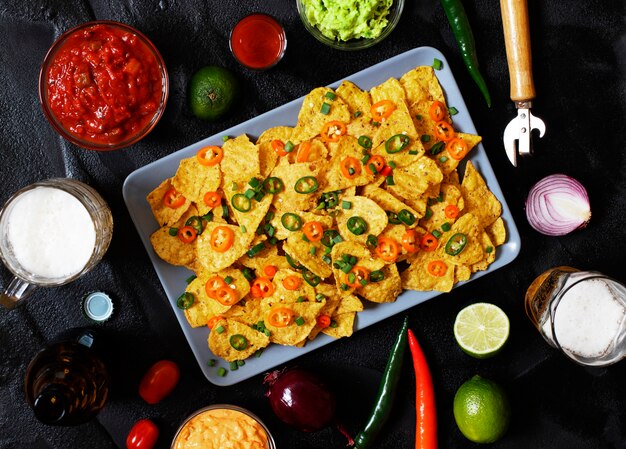 Mexikanische gelbe maistortillachips nachos mit jalapeno, guacamole, käsesoße und salsa. gläser mit bier, limette, chilischoten, kirschtomaten