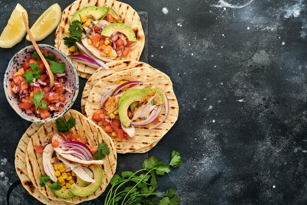 Mexikanische Tacos mit gegrilltem Hühnchen, Avocado, Maiskörnern, Tomaten, Zwiebeln, Koriander und Salsa am schwarzen Steintisch. Traditionelles mexikanisches und lateinamerikanisches Streetfood. Ansicht von oben.