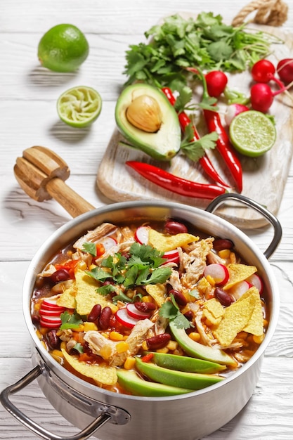 Mexikanische Tortillasuppe mit Pulled Chicken, Tomate, Mais, roten Kidneybohnen, Avocado, Rettich und Limette, Chili, Koriander, serviert auf einem Metalltopf auf weißem Holzhintergrund, Draufsicht, Nahaufnahme