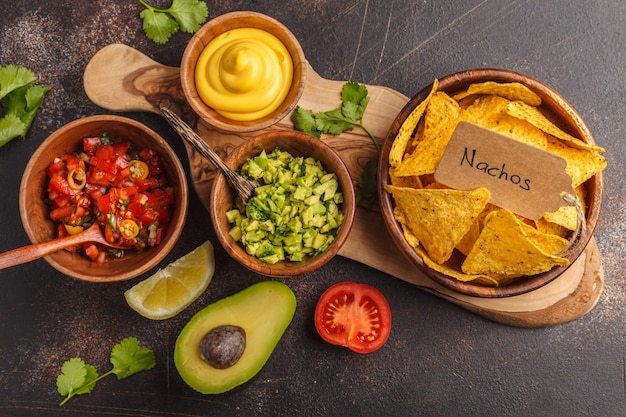 Mexikanisches Essen-Konzept. Nachos - gelbe Mais-Totopos-Chips mit verschiedenen Saucen in Holzschalen: Guacamole, Käsesauce, Pico del Gallo. Draufsicht, Lebensmittelhintergrund