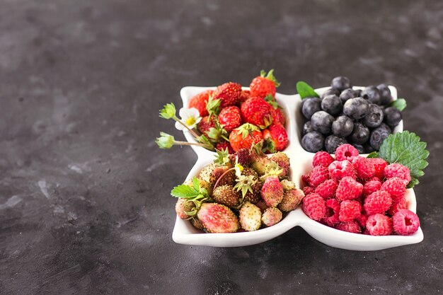 Una mezcla de bayas frescas en un plato blanco sobre un fondo gris oscuro, nutrición cruda natural