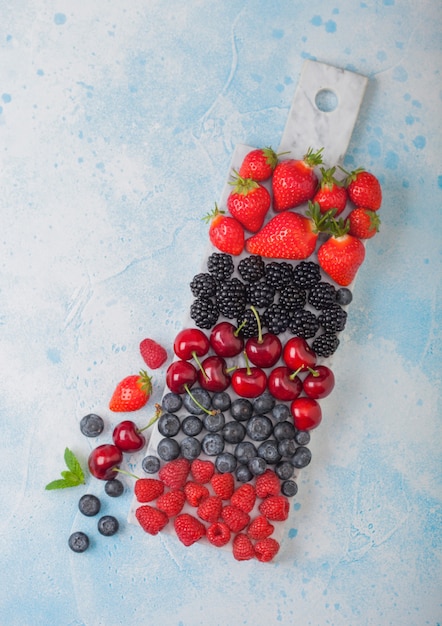 Mezcla de bayas de verano orgánico fresco en tablero de mármol blanco sobre fondo azul de la mesa de cocina. Frambuesas, fresas, arándanos, moras y cerezas. Vista superior