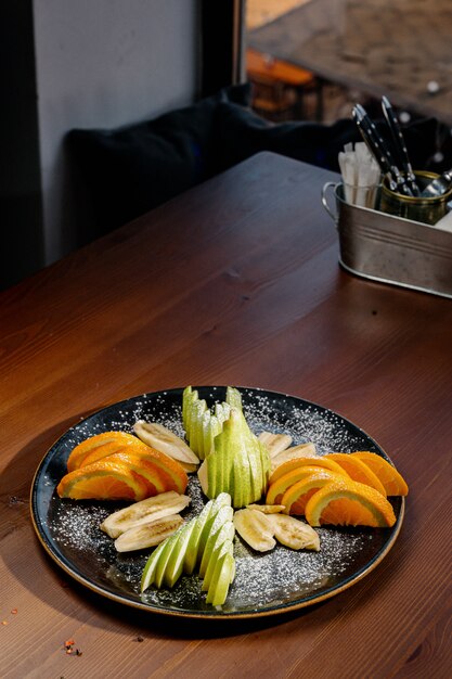 Mezcla de ensalada de frutas, postre de frutas: plátano en rodajas, mandarina, naranja, manzana. Frutas en rodajas en un plato negro. Un postre sabroso y saludable Vista superior