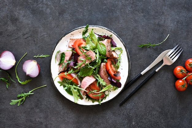 Mezclar la ensalada con los tomates y la carne mediana en un plato sobre un fondo oscuro.