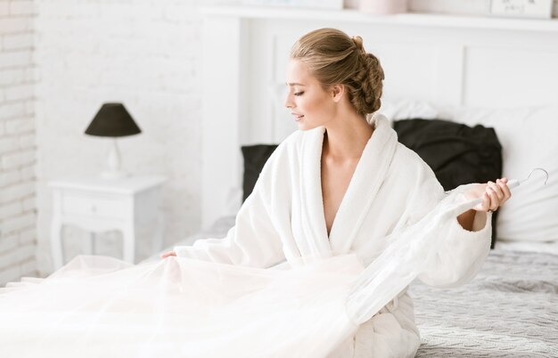 Mi precioso vestido. Encantadora novia joven alegre sentada en la cama blanca en casa y expresando ternura mientras se prepara para la ceremonia de la boda y sostiene el vestido de novia