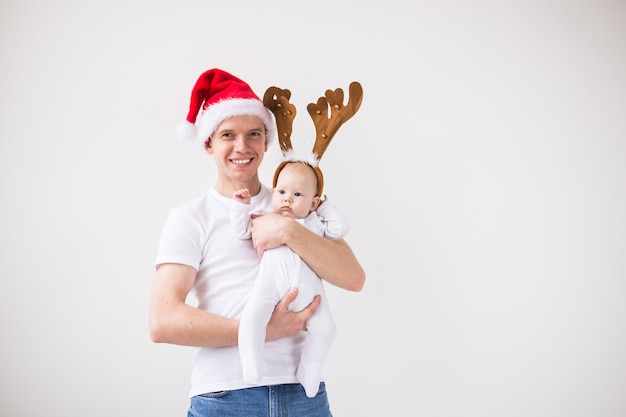 Mi primera Navidad. Papá con gorro de Papá Noel con su hija niña vistiendo cuernos de ciervo sobre fondo blanco con espacio de copia