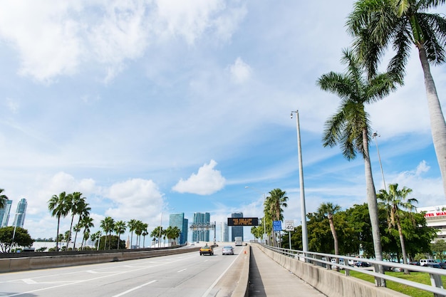 Miami, Estados Unidos - 30 de octubre de 2015: autopista o calzada con coches y rascacielos en el cielo azul nublado. Carretera con señales de tráfico para vehículos de transporte. Vallas publicitarias en edificios. Concepto de infraestructura pública.