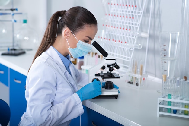 Microbiologia moderna Explorador concentrado bonito adulto sentado no laboratório em uma máscara de gaze trabalhando com o microscópio e conduzindo a pesquisa