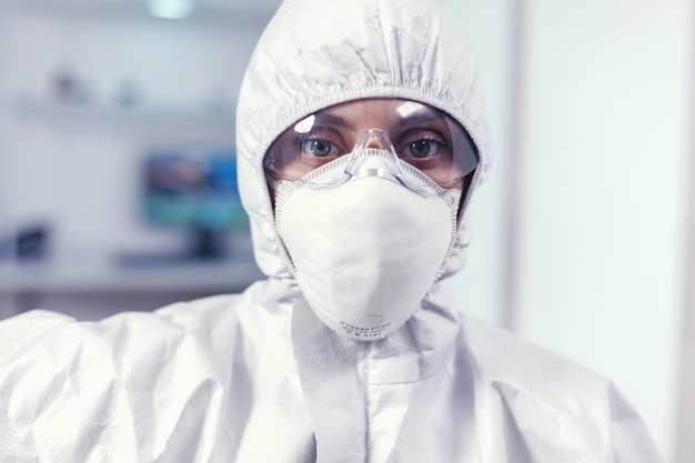 Microbiólogo agotado en mono sentado en el laboratorio mirando a la cámara. Científico de la mujer cansada en el laboratorio de biotecnología con traje protector durante la epidemia mundial.