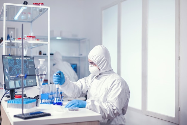 Microbiólogo vestido con mono con pipeta automática en laboratorio sanitario. Químico en laboratorio moderno haciendo investigación utilizando dispensador durante la epidemia mundial con covid-19.