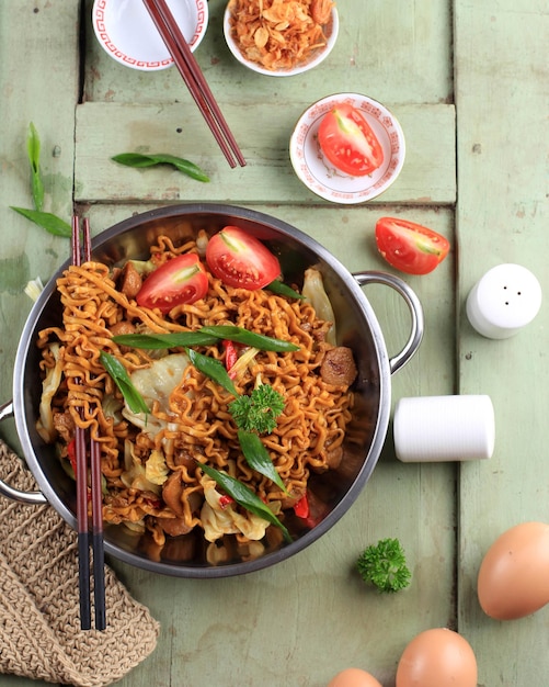 Mie Goreng oder Stir Fried Egg Noodles im chinesischen Stil ist traditionelles indonesisches Essen mit Huhn, Ei, Tomaten, Zwiebeln, Gemüse und süßer Sojasauce in einer weißen Platte, die auf einem weißen Holztisch serviert wird. Ansicht von oben.