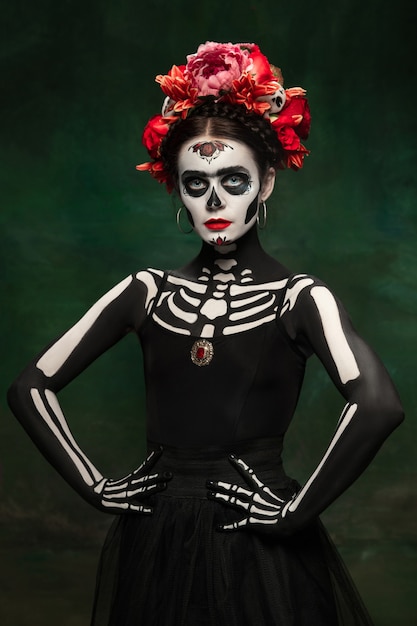 De miedo. Chica joven como Santa Muerte Santa muerte o Calavera de azúcar con maquillaje brillante. Retrato aislado sobre fondo de estudio verde oscuro con copyspace. Celebrando Halloween o Día de Muertos.