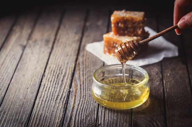 Miel que gotea del cucharón de miel en un tarro de vidrio con panal sobre fondo oscuro.