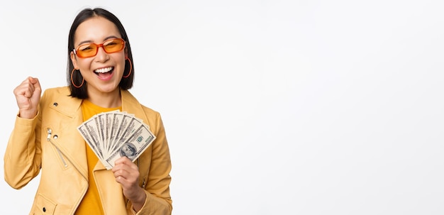 Mikrokredit- und Geldkonzept Stilvolle asiatische junge Frau mit Sonnenbrille, die glücklich lacht und Dollar-Bargeld hält, das über weißem Hintergrund steht