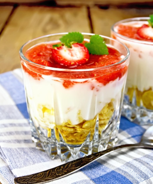 Milchdessert mit Erdbeercornflakes und Joghurt in einem Glas ein Löffel auf blauer Serviette auf einem Holzbretthintergrund