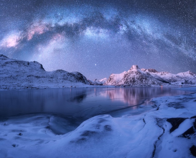 Milchstraße über gefrorener Meeresküste und schneebedeckten Bergen im Winter bei Nacht auf den Lofoten-Inseln Norwegen Arktische Landschaft mit blauem Sternenhimmel, Wasser, Eis, schneebedeckten Felsen, Milchstraße, Weltraum und Galaxie