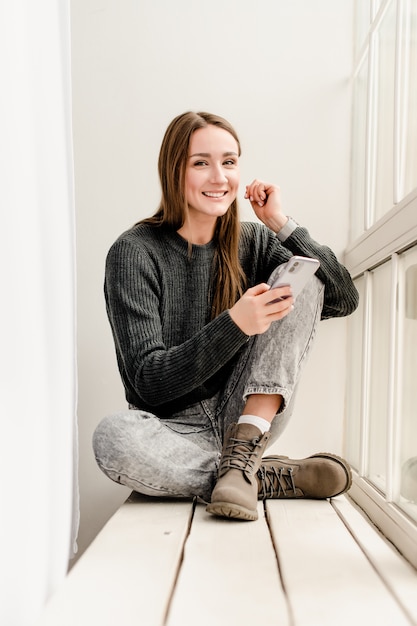 Milenar mulher sentada perto da janela com o telefone na mão