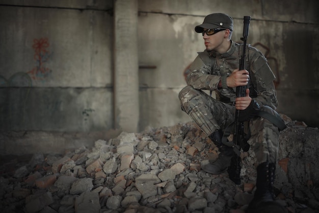 Un militar con una ametralladora en las manos con un uniforme de camuflaje gris con gorra y gafas tácticas se sienta en el suelo dentro de un gran edificio destruido