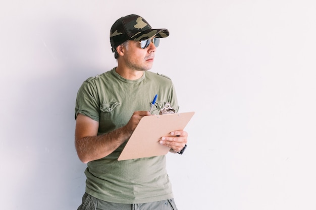Militar com boné camuflado e óculos escuros, escrevendo um relatório em uma pasta em uma parede branca