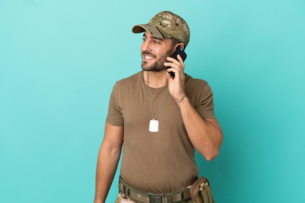 Militar com dog tag isolado em fundo azul, mantendo uma conversa com alguém no telefone celular