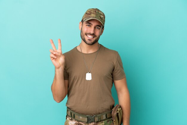 Militar com dog tag isolado em fundo azul, sorrindo e mostrando sinal de vitória