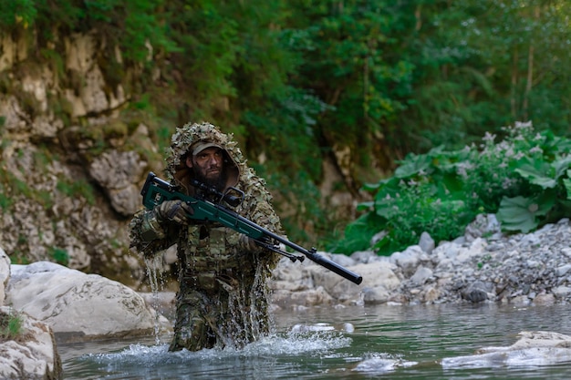 Un militar o un jugador de airsoft con un traje de camuflaje se esconde en el río y apunta con un rifle de francotirador hacia un lado o hacia el objetivo.
