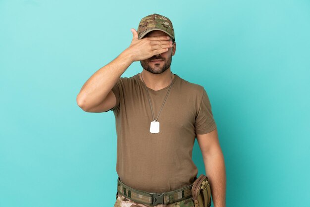 Militar con placa de identificación sobre aislado sobre fondo azul cubriendo los ojos con las manos. No quiero ver algo