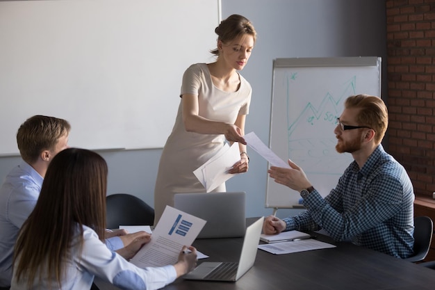 Millennial-Geschäftsfrau gibt den Teammitgliedern während einer Flipchart-Präsentation Unterlagen aus, weibliche Rednerin oder Coach teilt den Mitarbeitern Dokumente mit, die die Präsentantinnen in der Hand überlegen oder analysieren können.