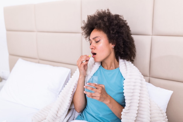 Millennial kranke kranke Frau, die Schmerzmittel Medizin nimmt, um Bauchschmerzen zu lindern, sitzen am Morgen auf dem Bett. Kranke Frau, die mit hohem Fieber im Bett liegt.