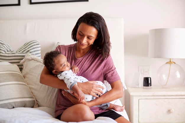 Millennial Mixed Race Mutter sitzt auf ihrem Bett und hält ihren drei Monate alten Sohn in den Armen