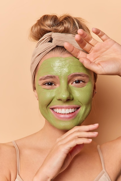 Mimar los procedimientos cosméticos para el cuidado del cuerpo y el concepto de spa. Mujer europea joven positiva aplica máscara de aguacate verde para el cuidado de la cara sonríe positivamente lleva diadema aislada sobre fondo beige.