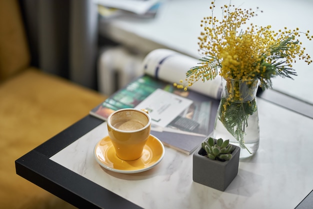 Mimose in einer Vase und Kaffee in einer gelben Tasse auf einem Tisch in einem Café oder Restaurant