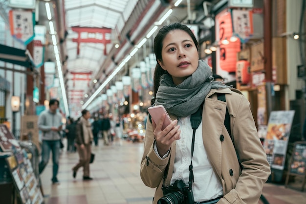 Minami Namba e Shinsaibashi. turista feminina em pé no mercado local e procurando loja de frutos do mar frescos durante o dia. jovem carregando câmera e segurando smartphone parece estilo de vida local japonês