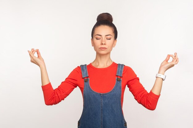 Mind-Balance-Atemtechnik Porträt eines ruhigen, stilvollen, hübschen Mädchens mit Haarknoten in Denim-Overalls, die Hände in Mudra-Geste halten und meditieren, während sie Yoga praktizieren, Studioaufnahme auf weißem Hintergrund