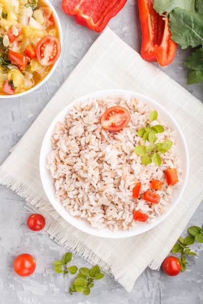 Mingau de arroz não polido com legumes cozidos e orégano em tigela branca sobre uma superfície de concreto cinza. Vista superior, close-up.