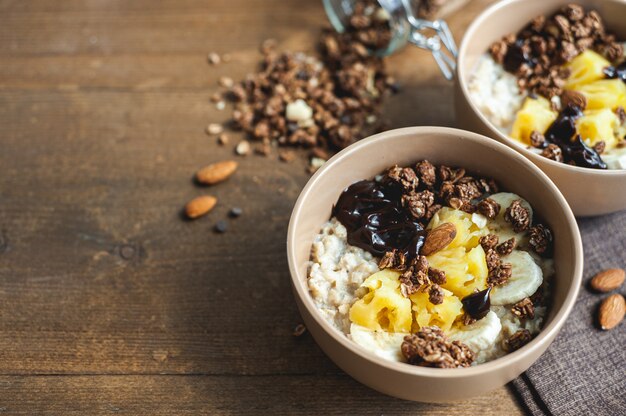 Mingau de aveia com abacaxi, banana e granola de chocolate em pratos bege em uma mesa de madeira marrom um farto ...