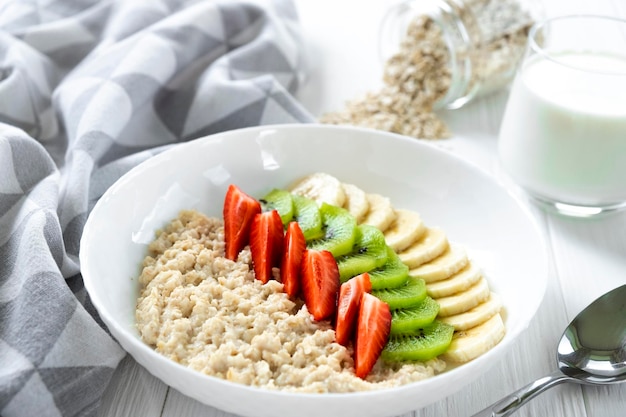 Mingau de aveia com frutas frescas morango banana kiwi em tigela branca com toalha de cozinha vidro de pote de leite com aveia e colher no fundo Receita de café da manhã saudável alimentação limpa