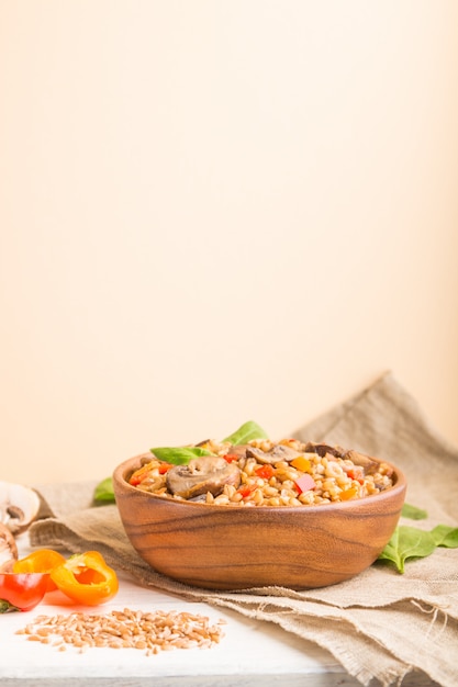 Mingau de espelta (trigo dinkel) com legumes e cogumelos em uma tigela de madeira sobre uma superfície branca. Vista lateral, foco seletivo, copie o espaço.