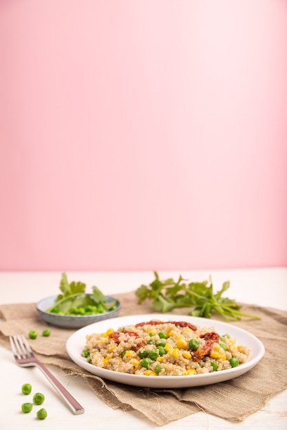 Mingau de quinua com ervilha, milho e tomates secos em prato de cerâmica sobre superfície branca e rosa pastel e tecido de linho