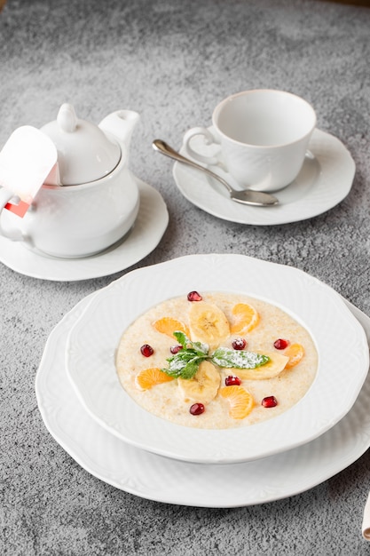 Mingau de sêmola ou aveia do café da manhã com bananas fatiadas em tigela branca isolada na mesa de pedra cinza. Comida caseira. Saboroso café da manhã. Foco seletivo. Foto vertical. Xícara e bule.