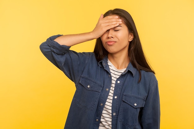 Minha culpa Retrato de menina triste e chateada em camisa jeans fazendo gesto de facepalm se culpando por evento esquecido erro estúpido memória ruim tiro de estúdio interno isolado em fundo amarelo