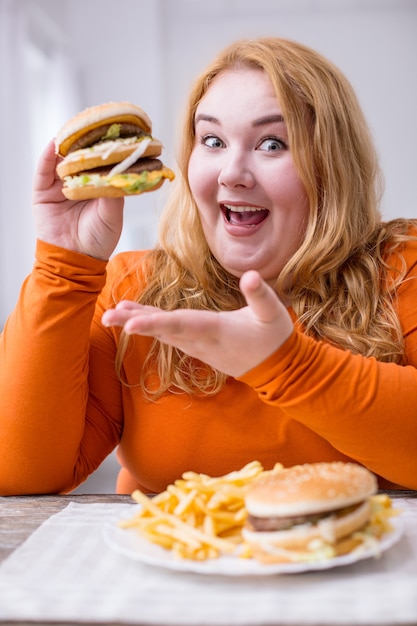 Minha felicidade. Mulher feliz e fraca com excesso de peso sentada à mesa comendo batatas fritas e sanduíches