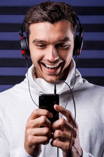 É minha música favorita! Jovem bonito usando fones de ouvido, segurando um MP3 player e olhando para ele com um sorriso, enquanto está de pé contra um fundo listrado