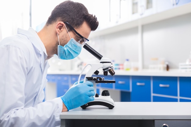 Minha pesquisa. moreno sério assistente de laboratório masculino posando em perfil enquanto olha no microscópio e estuda a amostra