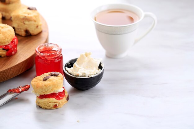 Mini bollos redondos con mermelada de fresa y crema cuajada y una taza de té, fondo de mármol blanco con espacio para copiar texto y receta