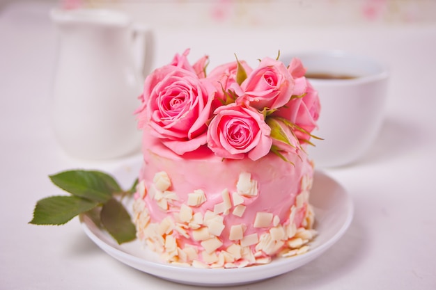 Mini bolo pequeno com esmalte rosa, lindas rosas, xícara de café, caixa de presente em cima da mesa branca.