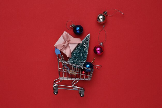 Mini carrito de compras con bolas de navidad mini árbol de navidad y caja de regalo sobre fondo rojo