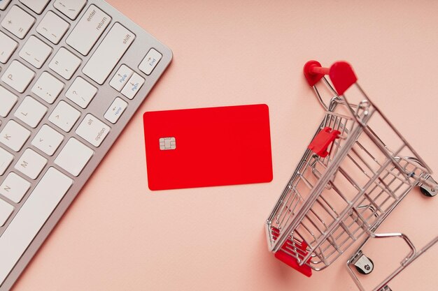 Mini carrito de compras y tarjeta de crédito roja con un teclado sobre fondo rosa Concepto de compras en línea