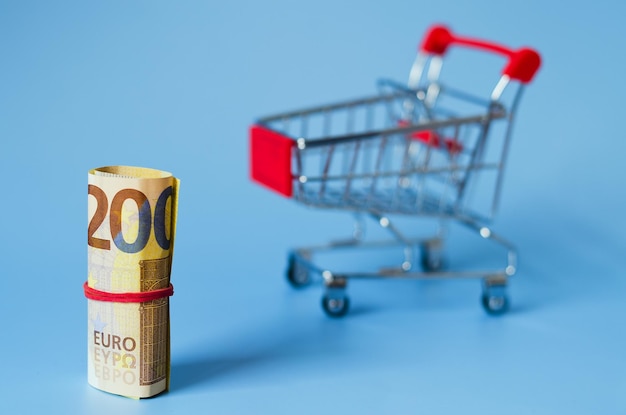 Mini carrito de supermercado con billetes en euros, dinero en una cesta, monedas. fondo azul.